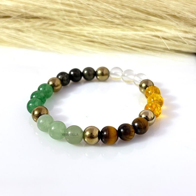 May include: A multi-coloured beaded bracelet with green, gold, clear, and brown beads. The bracelet is displayed on a white surface. The text "KRAFTILLA" is visible on the bracelet. The bracelet is made of various gemstones and metallic beads.