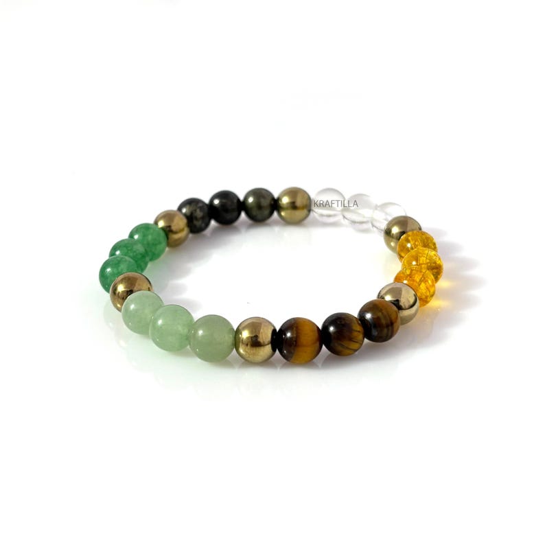 May include: A multi-coloured beaded bracelet with a variety of gemstones and metallic beads. The bracelet includes green, gold, brown, and clear beads. The beads are strung together on an elastic band. The brand name "KRAFTILLA" is visible.