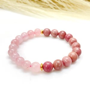 Könnte beinhalten: Ein Perlenarmband mit abwechselnd rosa und Rosenquarzperlen. Das Armband ist mit einem Gummiband versehen und hat goldfarbene Metallakzente. Die Perlen haben einen Durchmesser von ca. 1 cm.