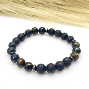 May include: A beaded bracelet featuring a combination of dark blue tiger eye, black lava rock, and dark gray hematite beads. The bracelet is strung on an elastic band and is displayed on a white surface.