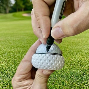 Peut inclure: Un marqueur noir trace une ligne sur une balle de golf blanche. La balle est maintenue dans un support en plastique blanc. L'arrière-plan est un parcours de golf vert.