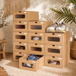 May include: A stack of brown cardboard shoe storage boxes with clear rectangular windows on the front panels. Each box features a small cutout handle at the top and a white label. Some boxes contain various pairs of athletic and casual footwear.