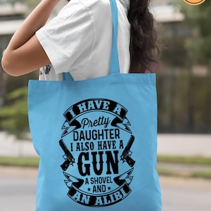 May include: A light blue tote bag with the text "I HAVE A Pretty DAUGHTER I ALSO HAVE A GUN A SHOVEL AND AN ALIBI" in a black and white design. The bag also features two gun illustrations. The words "EASY APPLICATION" and "PRINTED EXAMPLE" are also visible.