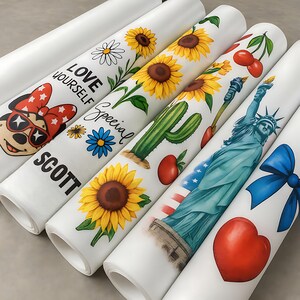 May include: Several rolls of white paper with colorful designs. One roll features a Minnie Mouse graphic, another has sunflowers and the words "LOVE YOURSELF" and "SCOTT". Other rolls display a cactus, cherries, the Statue of Liberty, and a blue bow.