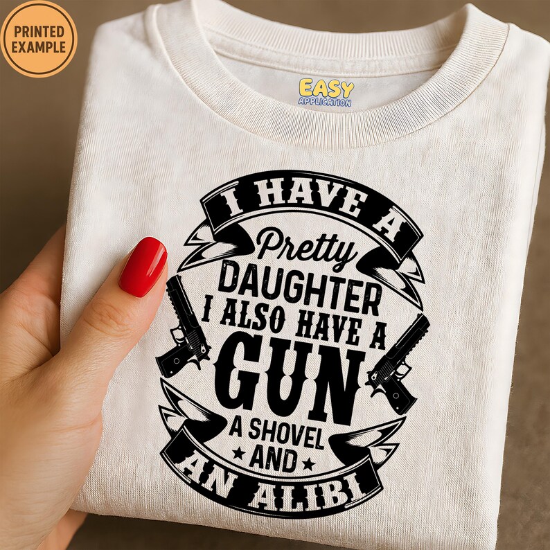 May include: Cream-colored t-shirt with a black graphic that reads "I HAVE A Pretty DAUGHTER I ALSO HAVE A GUN A SHOVEL AND AN ALIBI." The design includes two gun illustrations and a banner.