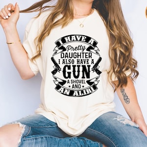 May include: Cream-colored t-shirt with a black graphic design. The design includes the text "I HAVE A Pretty DAUGHTER I ALSO HAVE A GUN A SHOVEL AND AN ALIBI" along with gun and shovel illustrations. A printed example label is visible.