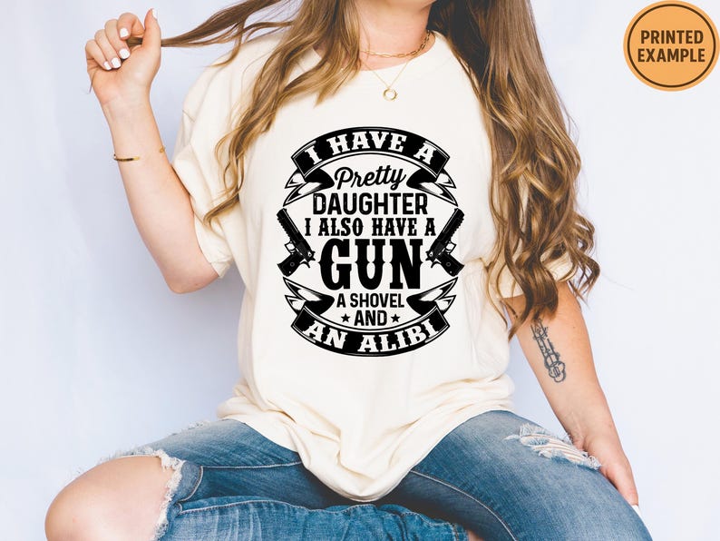 May include: Cream-colored t-shirt with a black graphic design. The design includes the text "I HAVE A Pretty DAUGHTER I ALSO HAVE A GUN A SHOVEL AND AN ALIBI" along with gun and shovel illustrations. A printed example label is visible.