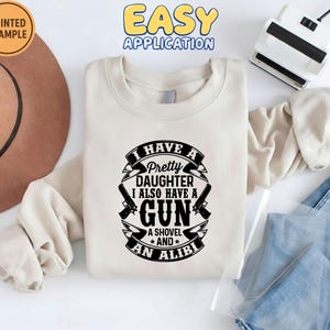 May include: Cream-colored sweatshirt with the text "I HAVE A Pretty DAUGHTER I ALSO HAVE A GUN A SHOVEL AND AN ALIBI" in black. The design includes gun and shovel graphics. The words "PRINTED EXAMPLE" and "EASY APPLICATION" are also visible.
