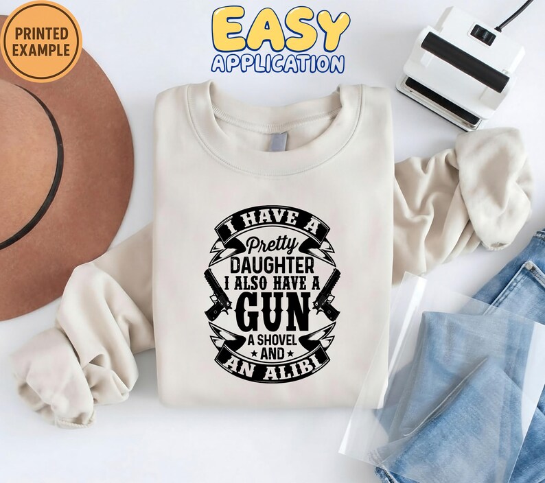 May include: Cream-colored sweatshirt with the text "I HAVE A Pretty DAUGHTER I ALSO HAVE A GUN A SHOVEL AND AN ALIBI" in black. The design includes gun and shovel graphics. The words "PRINTED EXAMPLE" and "EASY APPLICATION" are also visible.
