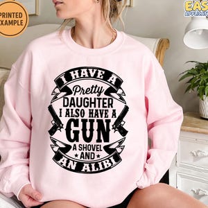 May include: A light pink sweatshirt with the text "I HAVE A Pretty DAUGHTER I ALSO HAVE A GUN A SHOVEL AND AN ALIBI" in black. The design includes two gun illustrations. The sweatshirt has long sleeves.