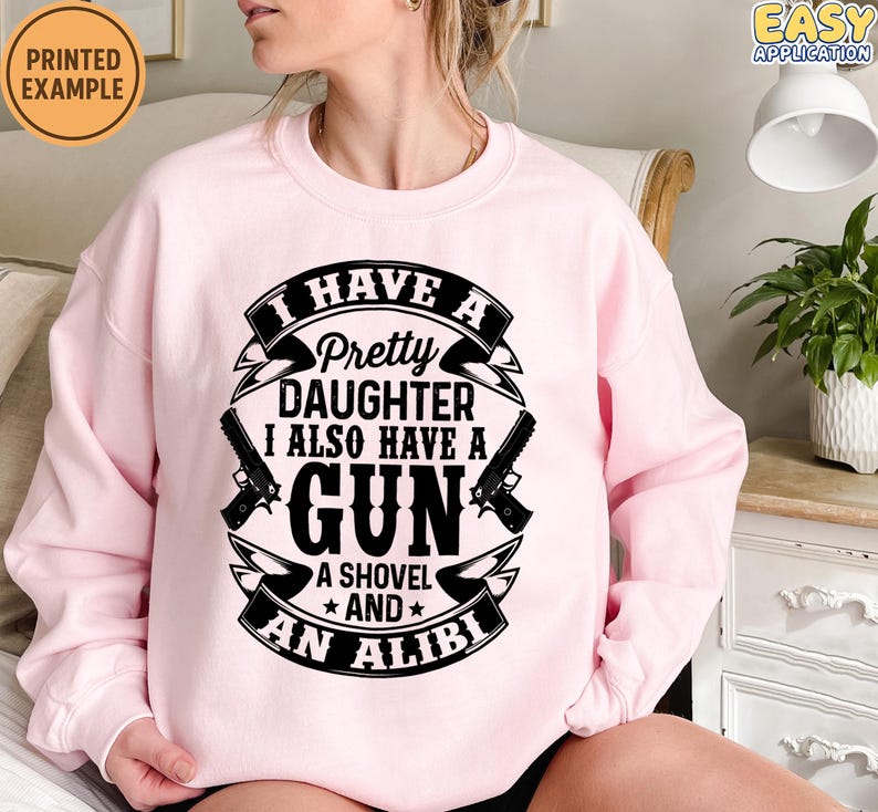 May include: A light pink sweatshirt with the text "I HAVE A Pretty DAUGHTER I ALSO HAVE A GUN A SHOVEL AND AN ALIBI" in black. The design includes two gun illustrations. The sweatshirt has long sleeves.