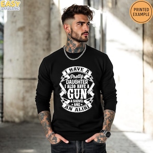 May include: Black long-sleeve sweatshirt with the text "I HAVE A Pretty DAUGHTER I ALSO HAVE A GUN A SHOVEL AND AN ALIBI" in white. The design includes two guns and a shovel graphic. The words "PRINTED EXAMPLE" are in a circle in the upper right corner.