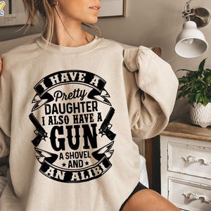 May include: Beige sweatshirt with a black graphic that reads "I HAVE A Pretty DAUGHTER I ALSO HAVE A GUN A SHOVEL AND AN ALIBI." The design includes two guns and a banner. The sweatshirt is a printed example.