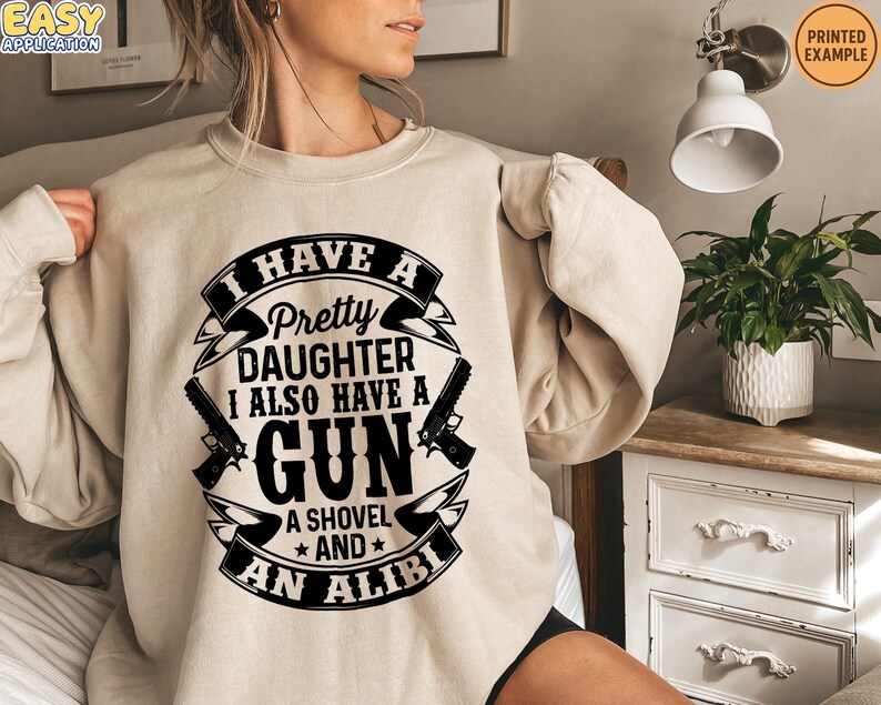 May include: Beige sweatshirt with a black graphic that reads "I HAVE A Pretty DAUGHTER I ALSO HAVE A GUN A SHOVEL AND AN ALIBI." The design includes two guns and a banner. The sweatshirt is a printed example.