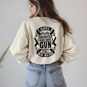 May include: Cream-colored sweatshirt with a black graphic on the back that reads "I HAVE A PRETTY DAUGHTER I ALSO HAVE A GUN A SHOVEL AND AN ALIBI". The sweatshirt has long sleeves and a relaxed fit.