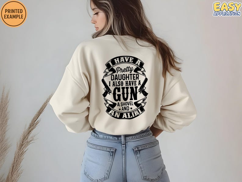 May include: Cream-colored sweatshirt with a black graphic on the back that reads "I HAVE A PRETTY DAUGHTER I ALSO HAVE A GUN A SHOVEL AND AN ALIBI". The sweatshirt has long sleeves and a relaxed fit.