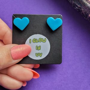 May include: A pair of blue heart-shaped earrings on a black card with the text "I GLOW IN UV".