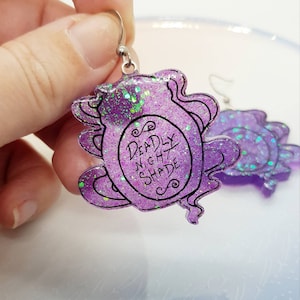 May include: Purple glitter resin earrings with a silver hook. The earrings are shaped like a bottle with a label that says "Deadly Nightshade".