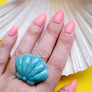 May include: A hand displaying a turquoise seashell ring with glitter, set against a large white seashell and a bright yellow background. The nails are painted a shimmery coral colour.