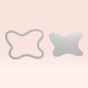 May include: Two grey, butterfly-shaped objects against a light pink background. One is a solid shape, and the other is an outline. The objects appear to be made of metal or a similar material. The shapes are symmetrical with rounded edges.