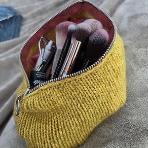 Puede incluir: Un neceser de maquillaje tejido amarillo con cremallera plateada, abierto para revelar brochas de maquillaje, un rizador de pestañas y otros productos de belleza. El neceser tiene un forro interior rosa.