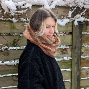 May include: A chunky knit infinity scarf featuring a variegated color palette of brown, tan, and orange tones. The scarf is worn around the neck and paired with a black textured fleece jacket that has a visible vertical seam along the sleeve.