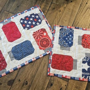 May include: Two patriotic quilted placemats. One is rectangular, the other square. Both feature red, white, and blue patterns including stars, polka dots, and a floral design. The borders are trimmed with a red, white, and blue checkered pattern.