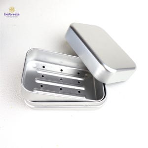 May include: A rectangular silver-colored metal soap dish with a removable lid. The base features three raised horizontal ridges with small drainage holes, and the lid is smooth and plain. The brand name herbreeze HANDMADE is visible.