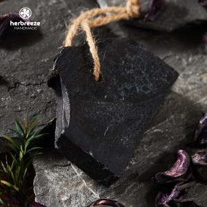 May include: A black, irregularly shaped soap bar with a textured surface, suspended by a natural twine loop. The soap is set against a dark, stone-like background, with the "herbreeze HANDMADE" logo in the upper left corner.