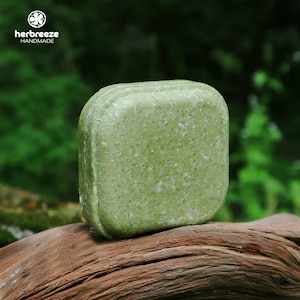 May include: A square, green shampoo bar with a textured surface. The bar is labeled "her breeze handmade" in white text. The bar is resting on a piece of driftwood, with a blurred green background.