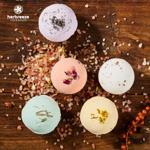 May include: Five colorful bath bombs with dried herbs and flower petals on a wooden surface. The bath bombs are light purple, peach, white, light blue, and yellow. The image includes the text "herbreeze HANDMADE".