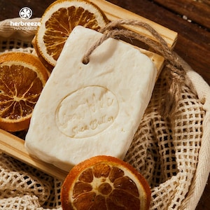 May include: A rectangular bar of handmade soap with a rope loop, featuring an oval imprint that reads "Happy Hippi Shea Butter." The soap is light beige and rests on a wooden tray with dried orange slices. The background includes a mesh bag.