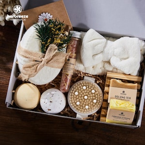 May include: A self-care gift box containing a white headband, bath bombs, a body brush, soap, a bath sponge, and bath salts. The box is lined with brown paper and decorated with dried flowers. The soap is labelled 'Oolong Tea'.