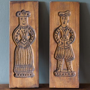 Pode incluir: Duas formas de madeira antigas com figuras esculpidas de um homem e uma mulher em trajes tradicionais. As figuras estão sobre um fundo castanho. As formas retangulares são provavelmente para assar.