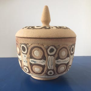 Puede incluir: Un tarro de cerámica beige con tapa. El tarro está decorado con un patrón repetitivo de círculos y óvalos en tonos marrones, beige y blancos. La tapa tiene un pomo cónico y un patrón similar en el borde. El tarro está sobre una superficie azul.