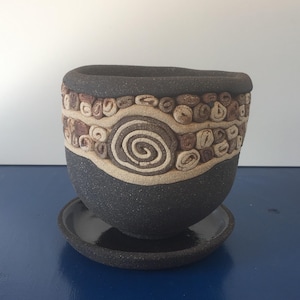 Op de afbeelding: Een handgemaakte, donkergrijze keramische plantenpot met bijpassende schotel. De pot heeft een getextureerd oppervlak en een decoratieve band met spiraal- en kiezelachtige ontwerpen in bruin- en beigetinten. Unieke, aardse esthetiek.