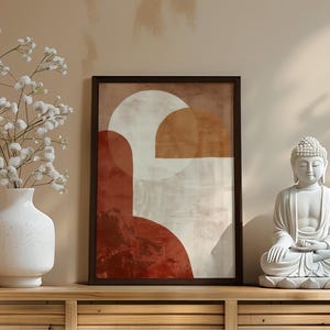 May include: A framed abstract art print with geometric shapes in shades of red, brown, and white. A white vase with white flowers and a white Buddha statue are on a wooden surface.