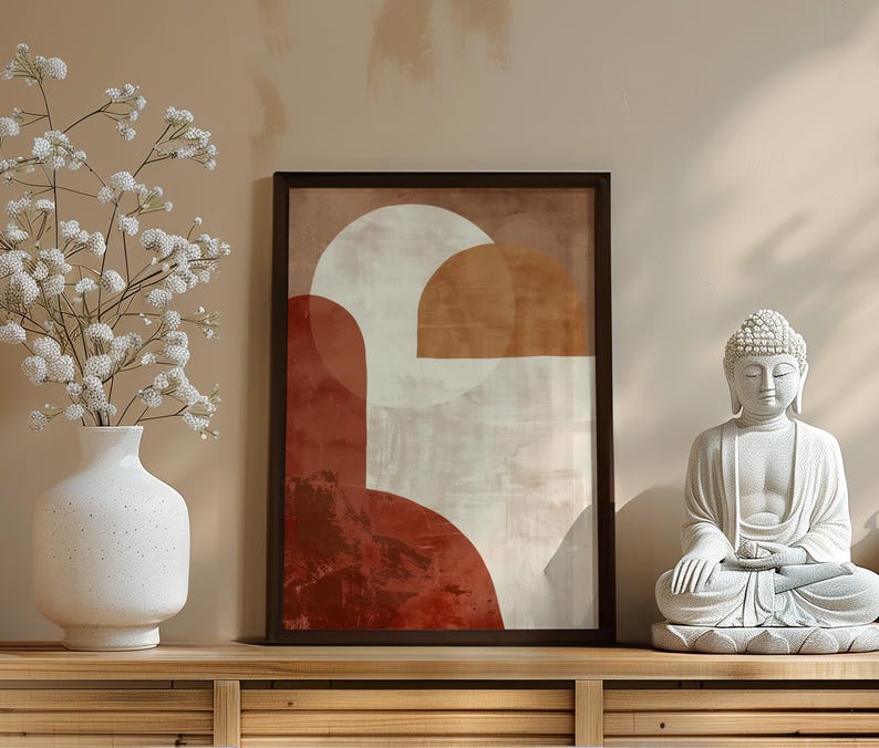 May include: A framed abstract art print with geometric shapes in shades of red, brown, and white. A white vase with white flowers and a white Buddha statue are on a wooden surface.