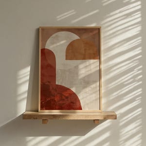 May include: Abstract art print in a wooden frame. The artwork features geometric shapes in shades of red, beige, and brown. The framed art is displayed on a wooden shelf against a white wall, with shadows from blinds.