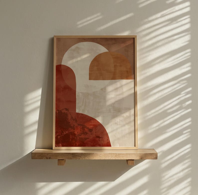 May include: Abstract art print in a wooden frame. The artwork features geometric shapes in shades of red, beige, and brown. The framed art is displayed on a wooden shelf against a white wall, with shadows from blinds.