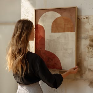 May include: Abstract art print in a wooden frame. The artwork features geometric shapes in shades of red, beige, and brown. The framed art is rectangular and is being held up against a textured wall.