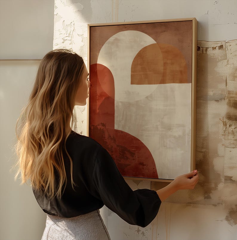 May include: Abstract art print in a wooden frame. The artwork features geometric shapes in shades of red, beige, and brown. The framed art is rectangular and is being held up against a textured wall.