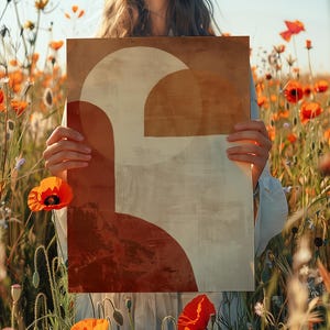 May include: An art print with a geometric design in brown, beige, and red tones is held in front of a field of orange poppies. The artwork features a large semi-circle and other abstract shapes. The print is held by a person. The image is set in a field of poppies.