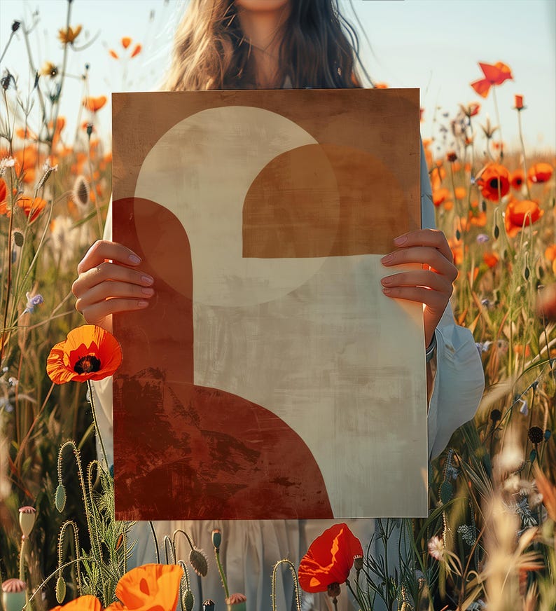 May include: An art print with a geometric design in brown, beige, and red tones is held in front of a field of orange poppies. The artwork features a large semi-circle and other abstract shapes. The print is held by a person. The image is set in a field of poppies.