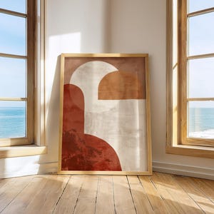 May include: A framed abstract art print with geometric shapes in red, beige, and brown. The artwork is displayed in a room with wooden floors and windows overlooking the sea. The frame is a light brown colour.