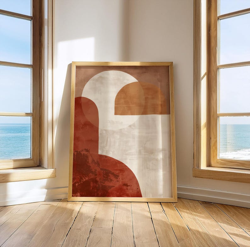 May include: A framed abstract art print with geometric shapes in red, beige, and brown. The artwork is displayed in a room with wooden floors and windows overlooking the sea. The frame is a light brown colour.