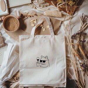 以下が含まれることがあります： サングラスをかけた猫がワイングラスを持っている黒い線画が描かれた、オフホワイトのキャンバスのトートバッグ。バッグには白いハンドルが2つ付いています。周りにはドライフラワーなどの装飾品があります。