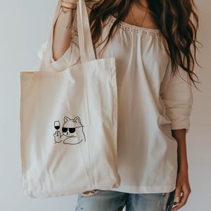 以下が含まれることがあります： サングラスをかけ、ワイングラスを持った猫の黒い線画が描かれた、クリーム色のトートバッグ。白いオフショルダーのトップスとブルージーンズを着た人がバッグを持っています。