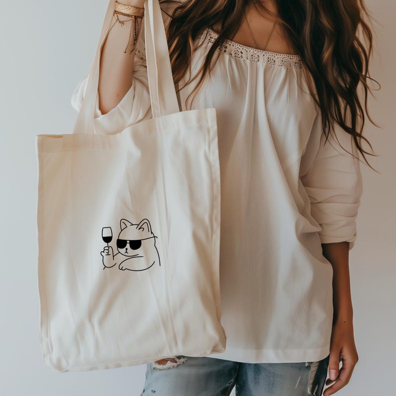 以下が含まれることがあります： サングラスをかけ、ワイングラスを持った猫の黒い線画が描かれた、クリーム色のトートバッグ。白いオフショルダーのトップスとブルージーンズを着た人がバッグを持っています。