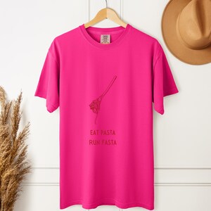 Peut inclure: T-shirt rose vif avec l'inscription "EAT PASTA RUN FASTA" en rouge foncé. Un dessin au trait de spaghettis sur une fourchette est au-dessus du texte. Le t-shirt est suspendu à un cintre en bois.